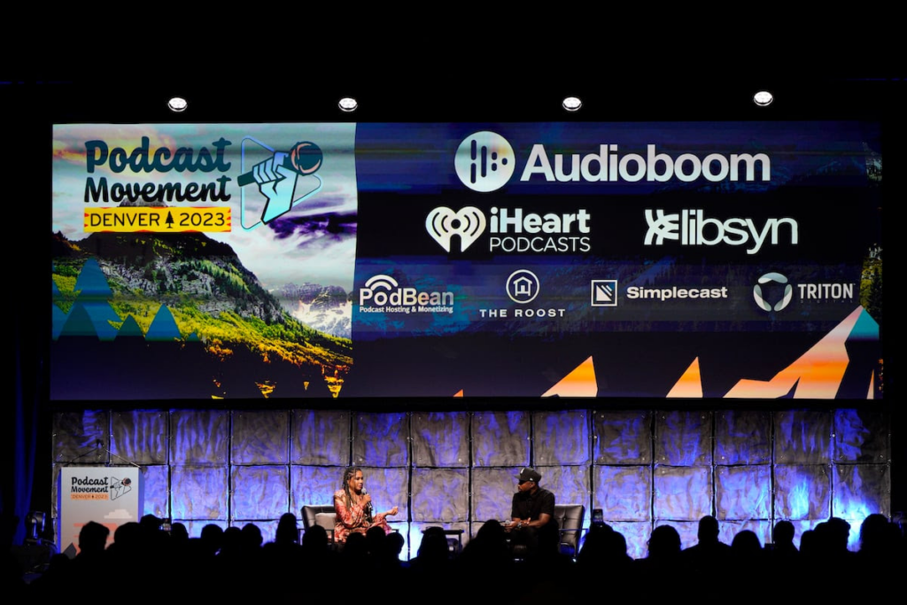 Image shows keynote stage from Podcast Movement 2023 Denver. There are 2 speakers sitting in the middle of the stage talking. 